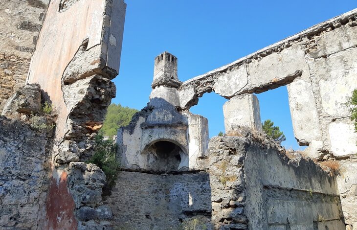 Kayaköy (Levissi)’de harabe halindeki bir evin iç duvarları, baca ve ocak kalıntısıyla birlikte açık gökyüzü altında görünüyor.