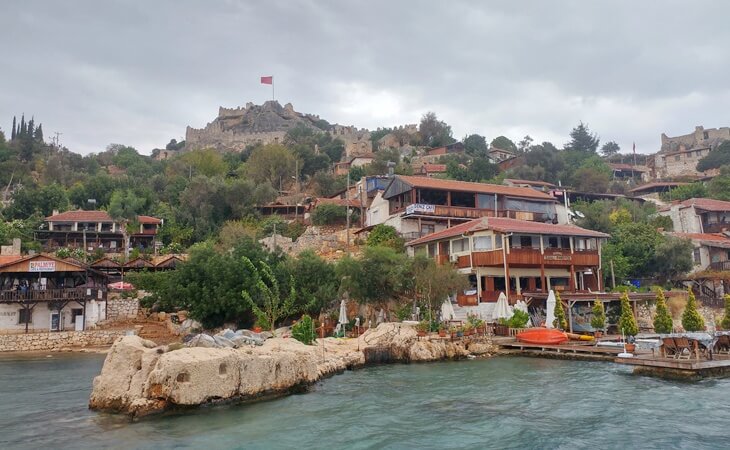 Bulutlu bir günde Kaleköy (Simena) sahili, deniz kenarında restoranlar ve arka planda Simena Kalesi