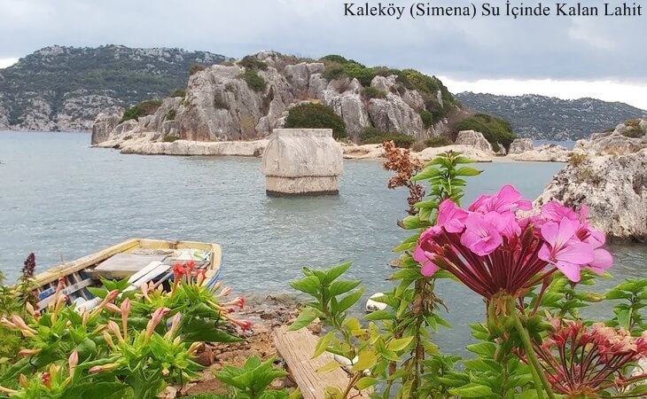 Kaleköy (Simena) kıyısında su içinde kalan antik lahit, ön planda çiçekler ve eski bir sandal