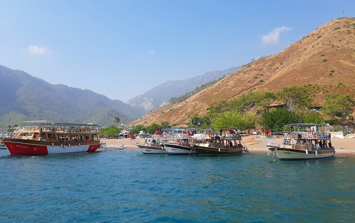 Adrasan Koyu sahilinde sıralanmış günübirlik tur tekneleri, arka planda ormanlık yamaçlar ve Akdeniz manzarası.