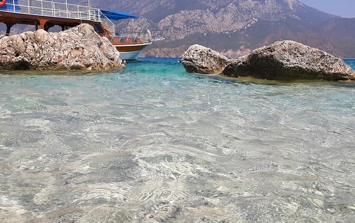 Antalya'nın Suluada kıyılarında berrak turkuaz sular ve kayalık sahil, arka planda demirlemiş bir tekneyle birlikte.