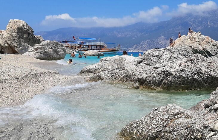 Antalya Suluada’da kayalık plajda denize giren insanlar, arkada demirli tekne ve dağ manzarası eşliğinde berrak deniz.