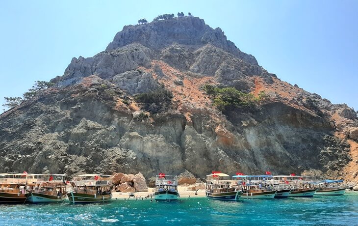 Antalya Suluada sahilinde sıralanmış tur tekneleri ve arka planda yükselen kayalık dağ manzarası.