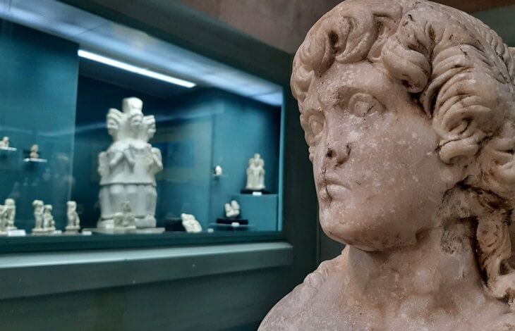 kutahya arkeoloji muzesi heykel bustu, mermer erkek figuru ve arka planda küçük arkeolojik eserlerin sergilendigi vitrin, roma dönemi buluntulari