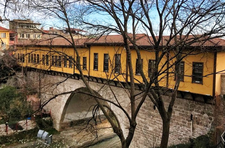 Bursa Irgandı K&ouml;pr&uuml;s&uuml;, ağa&ccedil; dalları arasından g&ouml;r&uuml;nen taş kemeri ve &uuml;zerindeki sarı renkli tarihi d&uuml;kk&acirc;nlarıyla nostaljik bir Osmanlı &ccedil;arşılı k&ouml;pr&uuml; manzarası