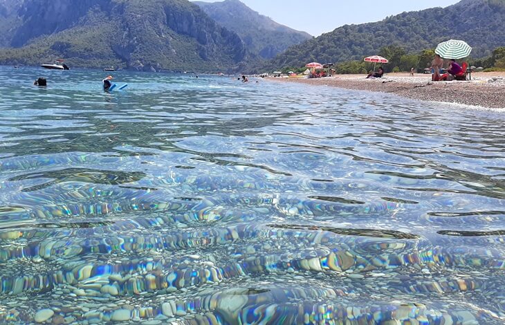 Olimpos Plajı’nda kıyıya yakın noktadan çekilmiş fotoğrafta berrak suyun altındaki çakıllar, kumsalda güneşlenen insanlar ve arka planda dağ silüetleri