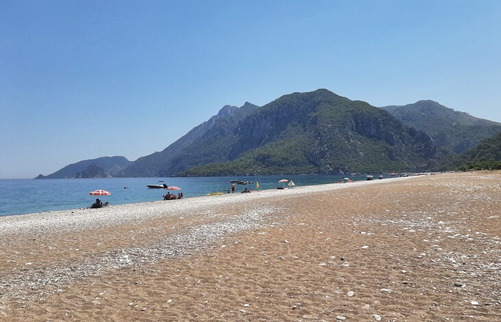Olimpos Plajı'nda geniş kumsal, denizde yüzen tekneler ve arka planda yemyeşil dağ manzarası