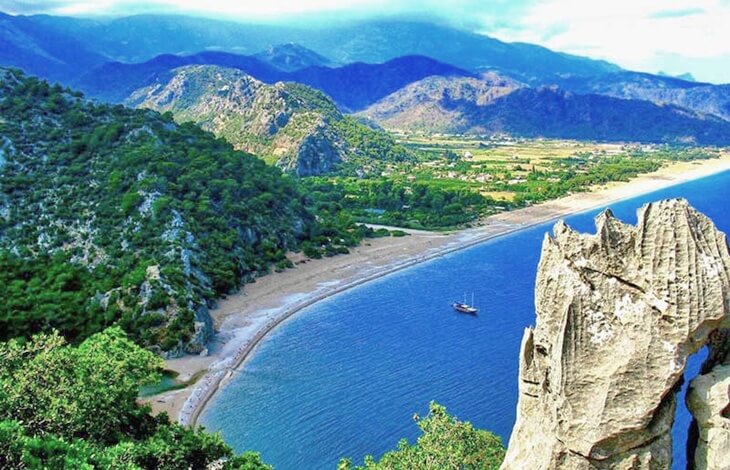 Olimpos Plajı'nın yukarıdan görünüşü; yeşil dağlar, geniş kumsal, masmavi deniz ve kıyıya demirlemiş bir tekneyle doğal bir manzara