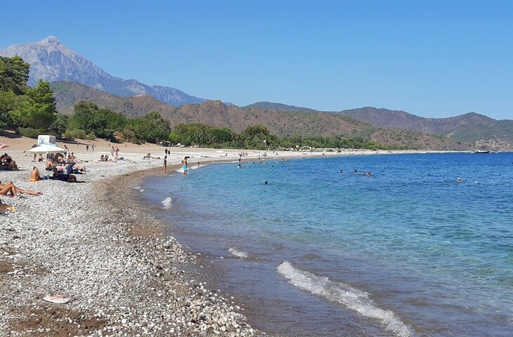 Olimpos Plajı'nda denize giren insanlar, çakıllı sahil ve arka planda Tahtalı Dağı manzarası