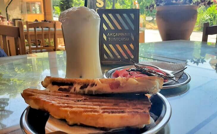 Akçapınar Tostçusu’nda servis edilen bol malzemeli tost, yanında ayran ve domates-biber garnitürüyle birlikte sunuluyor.
