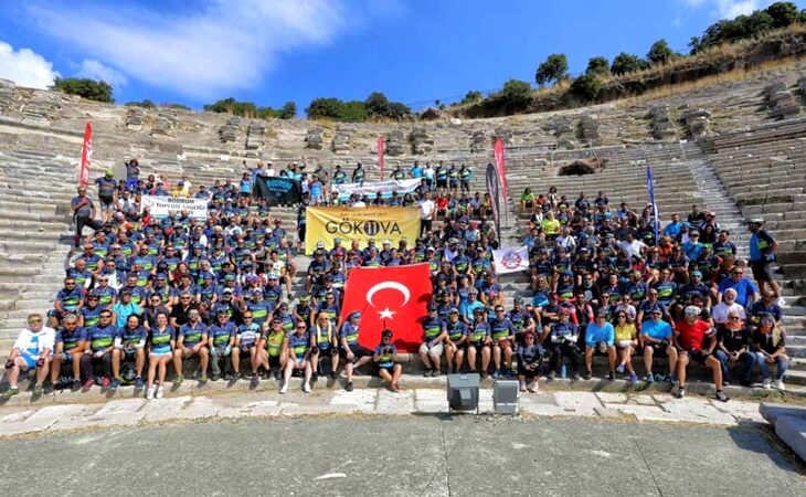 Tarihi bir antik tiyatroda toplanmış Gökova Bisiklet Turu katılımcıları, takım formalarıyla birlikte Türk bayrağı ve Gökova pankartı tutuyor.
