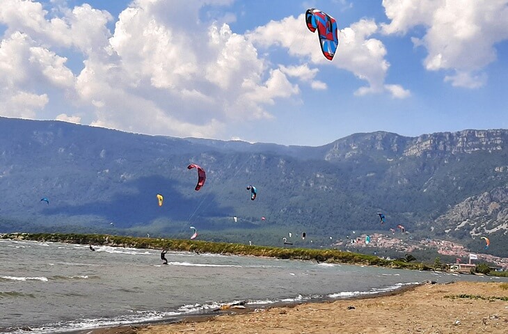 Akyaka denice akıllara rüzgar sörfü yapan kitesurfçüler geliyor; gökyüzünde rengarenk uçurtmalar ve arka planda dağ manzarası.