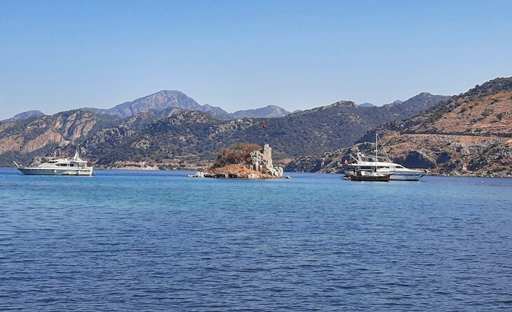 Marmaris Selimiye a&ccedil;ıklarında, ortasında bayraklı k&uuml;&ccedil;&uuml;k bir tarihi yapının bulunduğu kayalık ada ve &ccedil;evresinde demirlemiş tekneler.