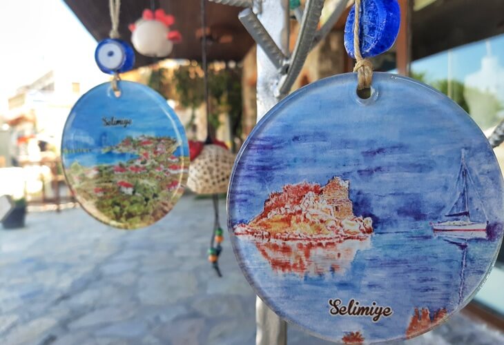 Marmaris Selimiye&rsquo;de el yapımı seramik s&uuml;sler, &uuml;zerinde Selimiye manzarası ve nazar boncuğu detaylarıyla bir d&uuml;kkan &ouml;n&uuml;nde asılı duruyor.