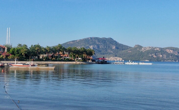 Marmaris Selimiye&rsquo;de sabah saatlerinde deniz kıyısı, palmiyelerle &ccedil;evrili evler, iskele ve dağ manzarası eşliğinde sakin bir koy g&ouml;r&uuml;n&uuml;m&uuml;.