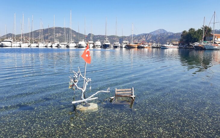 Selimiye&rsquo;de deniz i&ccedil;inde yerleştirilmiş bir sandalye, beyaz boyalı k&uuml;&ccedil;&uuml;k bir dal ve T&uuml;rk bayrağı; arka planda yelkenliler ve dağ manzarası.