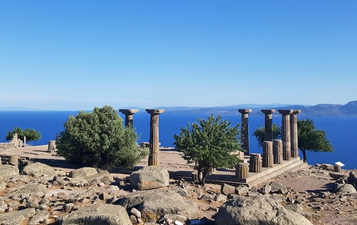 Assos Akropol&uuml;&rsquo;nde Athena Tapınağı&rsquo;nın Dor d&uuml;zeni s&uuml;tun kalıntıları; &ouml;n planda kaya bloklar ve zeytin ağa&ccedil;ları, arka planda masmavi Ege ve Midilli silueti.