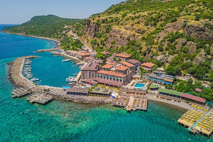 Assos Antik Limanı ve denize sıfır otellerin g&ouml;r&uuml;ld&uuml;ğ&uuml;, kayalık yama&ccedil;ların &ccedil;evrelediği turkuaz koyun havadan g&ouml;r&uuml;nt&uuml;s&uuml;.