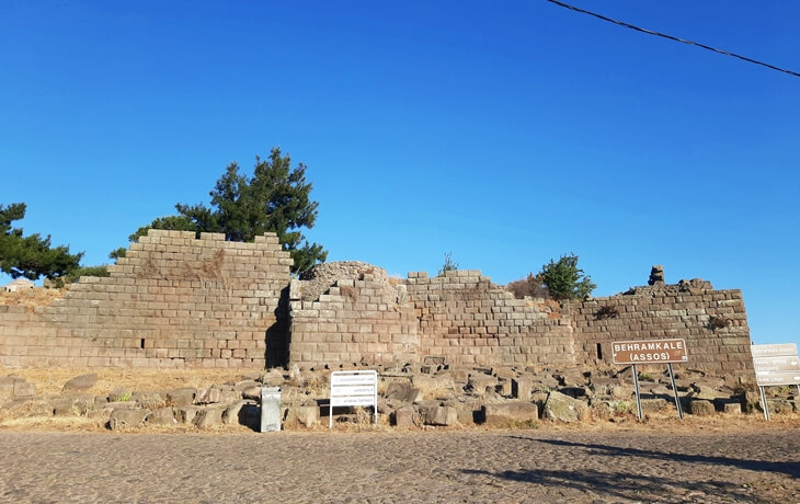 Assos/Behramkale girişindeki antik sur kalıntıları; kesme taş bloklar, &ouml;n&uuml;nde &ldquo;Behramkale (Assos)&rdquo; tabelası ve uyarı panoları.