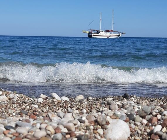 Olympos Plajı'nda çakıllı kıyı, kıyıya vuran küçük dalgalar ve açıkta duran bir yelkenli tekne