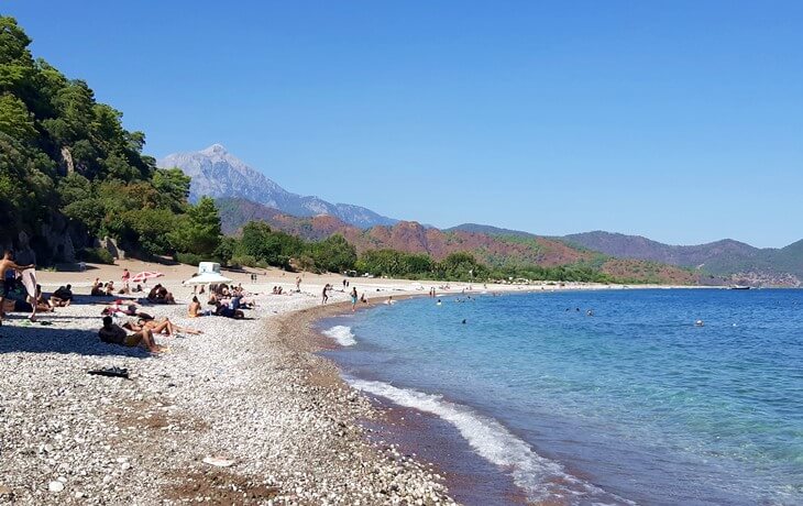 Olympos Plajı'nda güneşlenen insanlar, berrak deniz ve arka planda yükselen Tahtalı (Olympos) Dağı manzarası