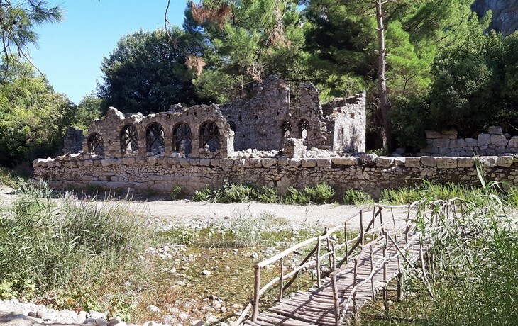 Olympos Antik Kenti'nde Olympos Çayı kıyısında yer alan taş rıhtım kalıntıları ve ön planda ahşap bir köprü