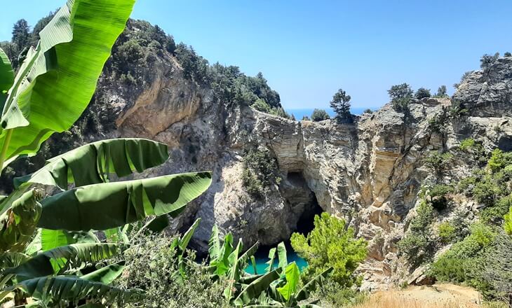 Gazipaşa Delikdeniz Kral Koyu, ön planda muz yaprakları ve yeşillikler arasında, kayalıklar ve doğal mağarayla birlikte görülüyor.