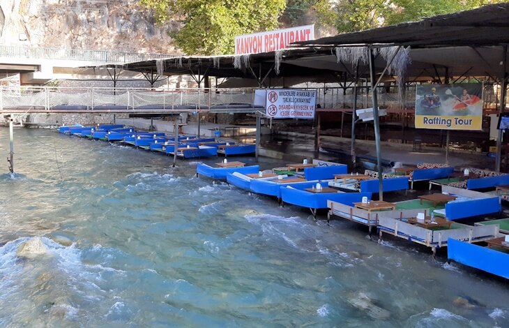 Saklıkent Kanyonu girişinde akan su üstüne kurulmuş minderli restoran masaları ve rafting turu afişi